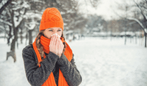 Winter Wellness For Teachers Shown Through A Teacher Standing Outside In The Snow, Wearing A Hat And Scarf, Blowing Her Nose.
