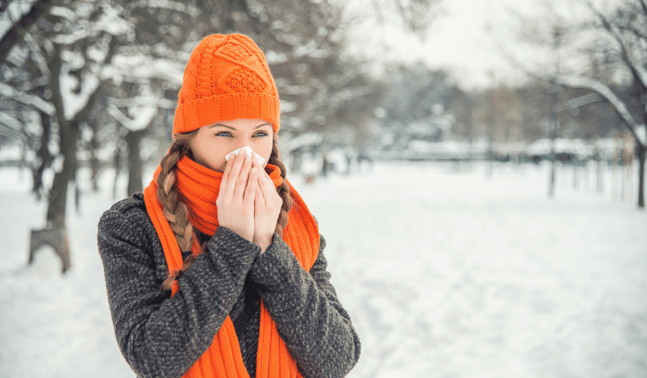 Winter wellness for teachers shown through a teacher standing outside in the snow, wearing a hat and scarf, blowing her nose.